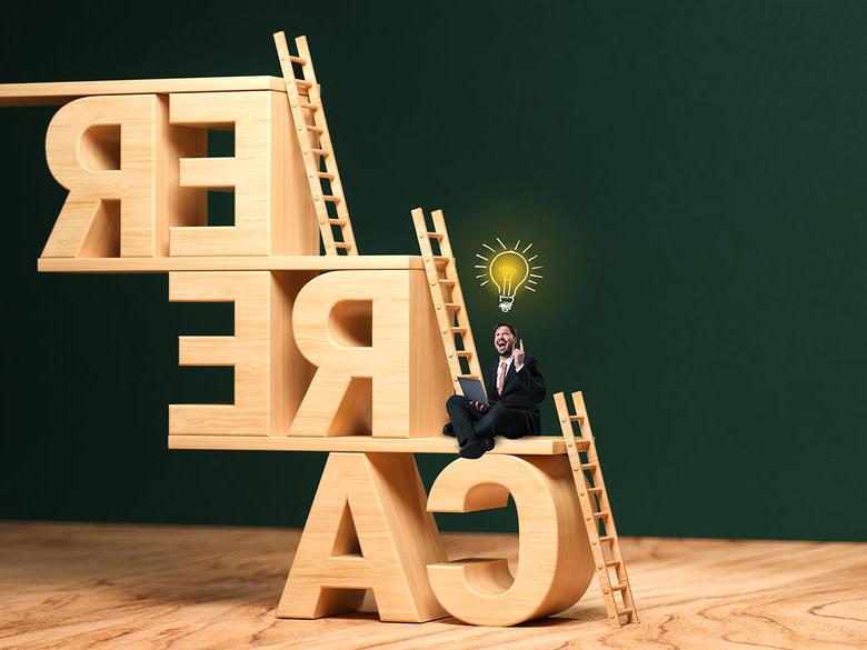 A man in a suit sits on large, wooden block letters spelling CAREER, holding a laptop, with ladders leaning against the letters and a lightbulb above his head.